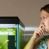 A woman stands next to a fish tank that smells bad, with a distorted expression.